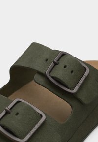 Πράσινο σατινέ σανδάλι με δύο ρυθμιζόμενες αγκράφες από σκοτεινό μέταλλο και περιβαλλόμενο ταμπά πέλμα ορατό κάτω από τους ιμάντες.