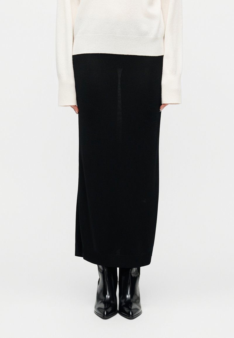 Άτομο που φοράει λευκό πουλόβερ με μακριά μανίκια, μαύρη φούστα μέχρι τον αστράγαλο και γυαλιστερά μαύρα μυτερά μποτάκια, στέκεται μπροστά σε λευκό φόντο.