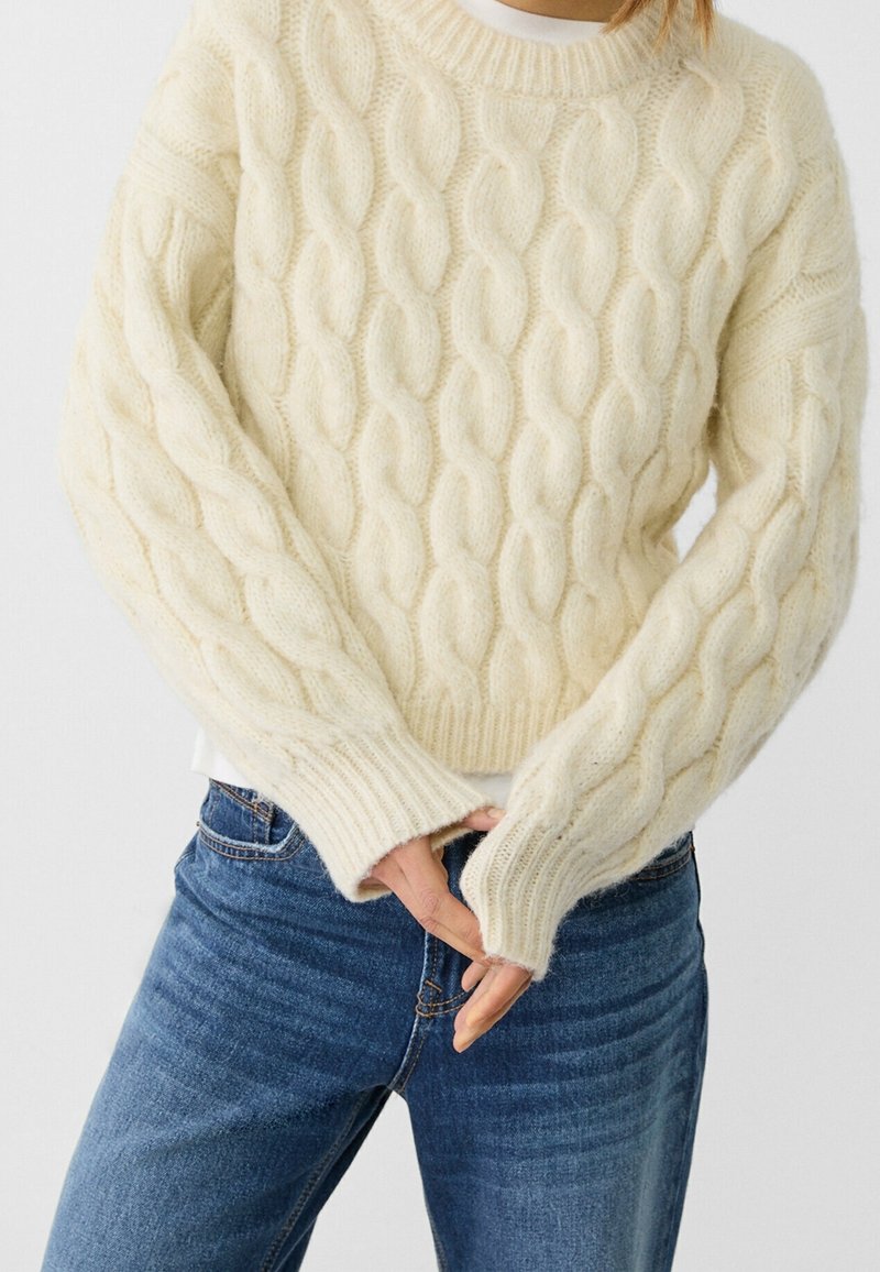 Personne portant un pull en maille torsadée de couleur crème et un jean bleu, les mains jointes devant et une chemise blanche dépassant dessous.