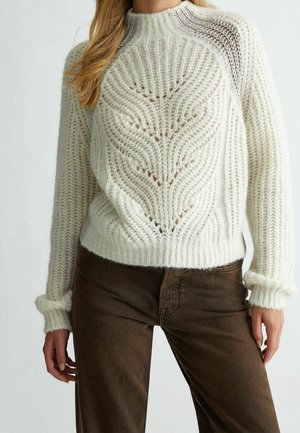 Femme portant un pull en maille blanche avec un motif chevron texturé et un pantalon marron, présentée du cou aux hanches sur un fond neutre.