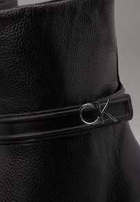 Černé kožené kotníkové boty s texturovaným povrchem, elegantním designem a kovovým stříbrným akcentem CK na černém pásku.