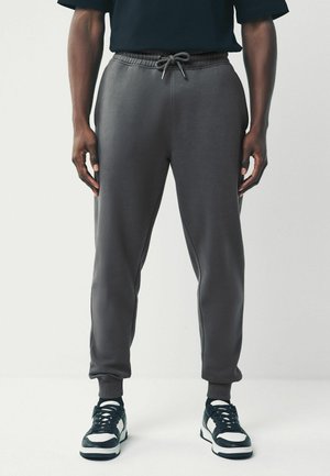 Pantalons de jogging gris avec taille élastique et cordon de serrage. Tissu doux, design fuselé, poches latérales, associés à des baskets noires et blanches.