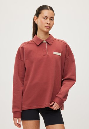 Ung kvinde med langt hår i en hestehale iført en rustfarvet langærmet sweatshirt med krave og sorte shorts, stående mod en ensfarvet baggrund.