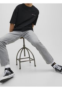 Camiseta negra de manga corta con un logo, combinada con jeans de pierna recta en gris claro y zapatillas negras con detalles en blanco, sentado en un taburete.