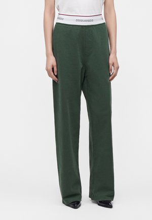 Personne portant un pantalon large vert foncé et un haut blanc avec une ceinture de marque visible, debout devant un fond clair uni.