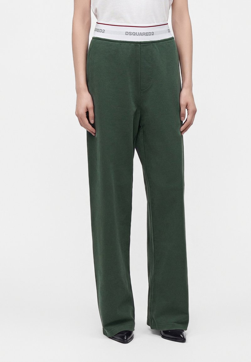 Personne portant un pantalon large vert foncé et un haut blanc avec une ceinture de marque visible, debout devant un fond clair uni.