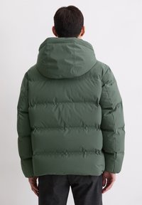 Veste compressible verte avec capuche, dotée d'un design matelassé et de coutures horizontales. Sa texture lisse et sa coupe décontractée sont mises en avant.