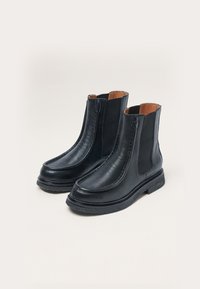 Botas de tobillo de cuero negro con paneles laterales elásticos, puntera redonda y suela de goma negra texturizada. Presenta un diseño sutilmente cosido.