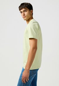 Ljusgrön kortärmad T-shirt, rund halsringning, i bomull, figursydd form. Bärs tillsammans med blå denimjeans, sidorprofil vy.