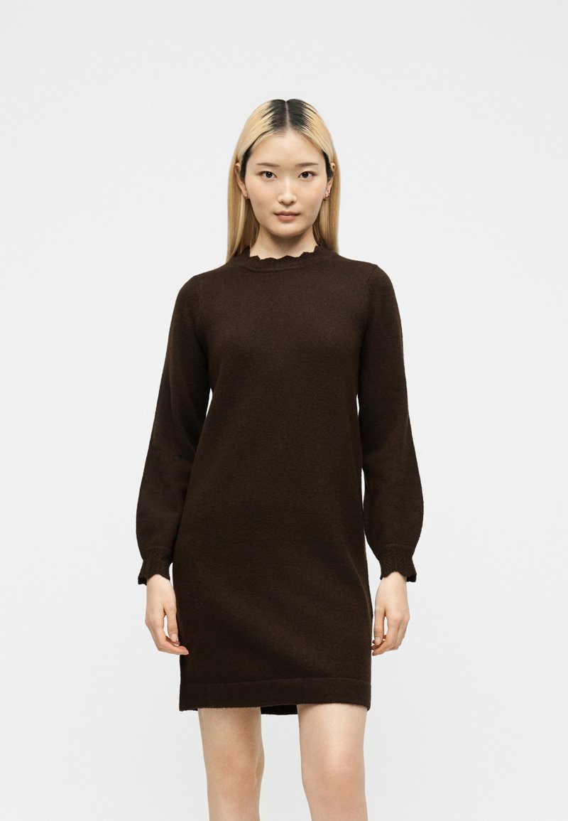 Femme aux cheveux blonds et raides portant une robe pull marron foncé à manches longues, longueur genou, debout devant un fond uni.
