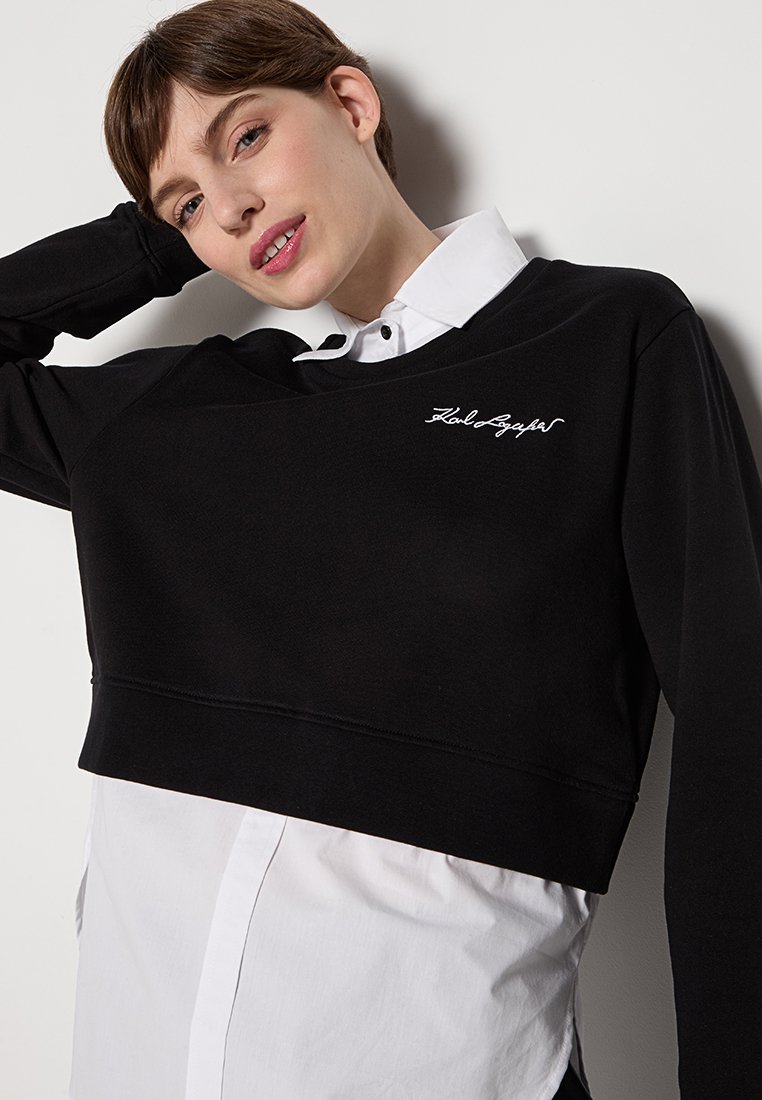Sweatshirt noir court à manches longues, arborant un logo brodé blanc. Porté par-dessus une chemise blanche à col, mettant en valeur un design superposé.