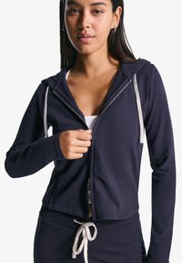 A young woman with long dark hair zips up a navy hoodie, revealing a white top underneath, set against a plain white background.