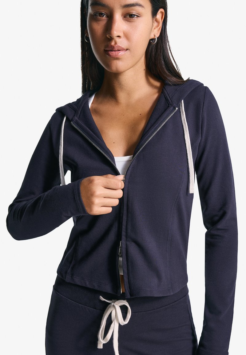 A young woman with long dark hair zips up a navy hoodie, revealing a white top underneath, set against a plain white background.