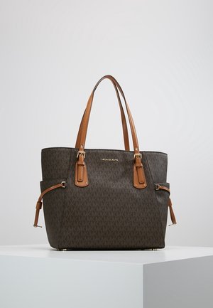 Brązowa torba typu tote Michael Kors z jasnobrązowymi skórzanymi rączkami, bocznymi kieszeniami i złotymi akcentami na białym postumencie na jednolitym tle.
