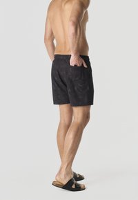 Uomo senza maglietta che indossa pantaloncini da nuoto neri con motivi e sandali neri, in piedi con una mano nella tasca posteriore contro uno sfondo chiaro.