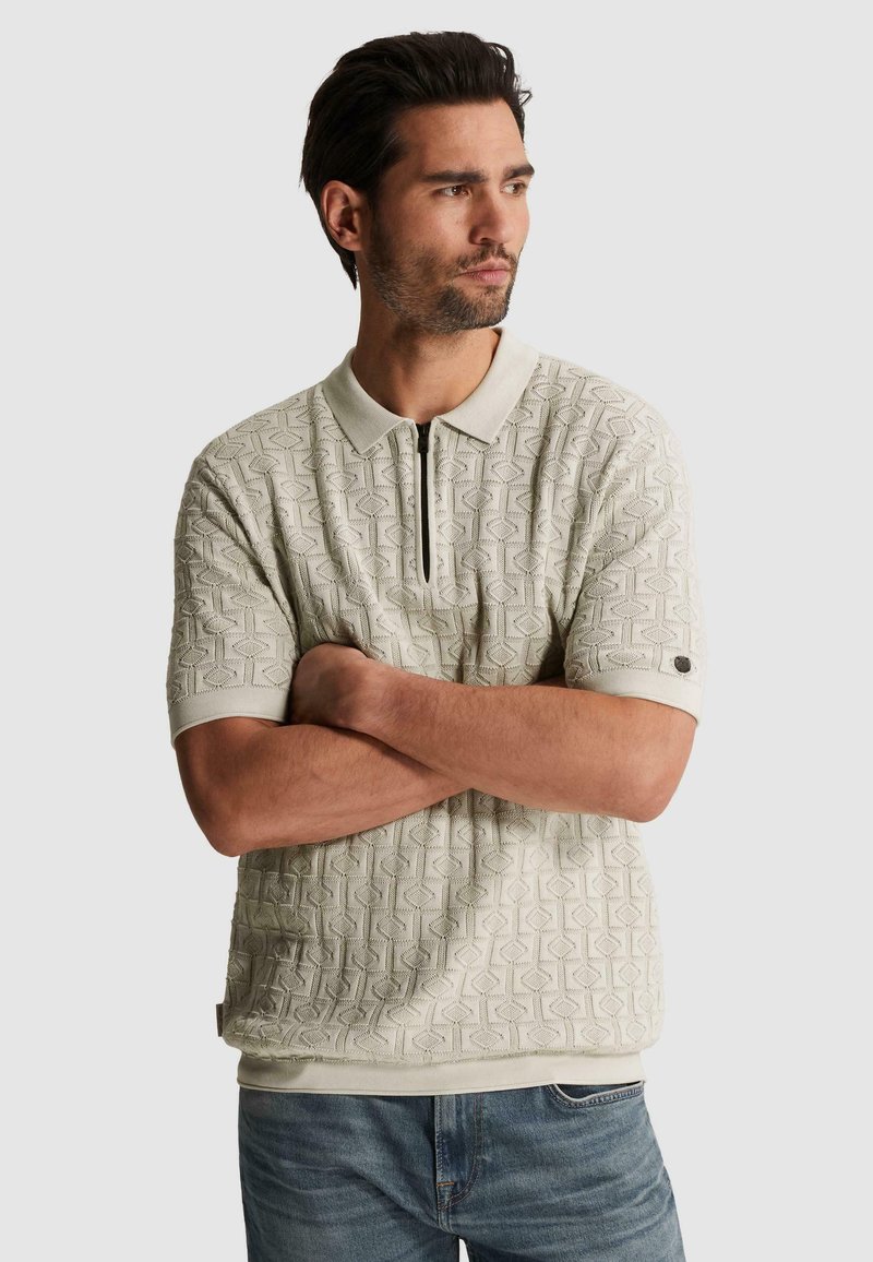 Homme avec les bras croisés portant un polo beige texturé à manches courtes et un jean bleu, regardant sur le côté sur un fond uni.