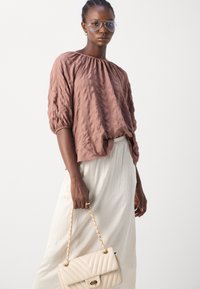 Blushfarbene, strukturierte Bluse mit Puffärmeln, kombiniert mit einem fließenden weißen Rock. Das Model hält eine cremefarbene, gesteppten Handtasche mit goldener Kette.