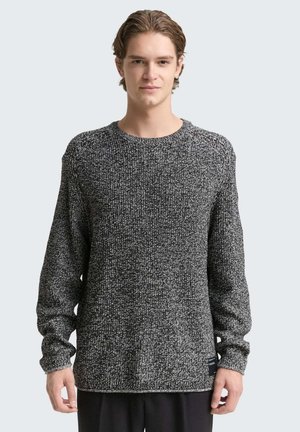 Junger Mann mit kurzen Haaren, der einen langärmligen grau-schwarz melierten Strickpullover trägt und vor einem einfarbigen Hintergrund steht.