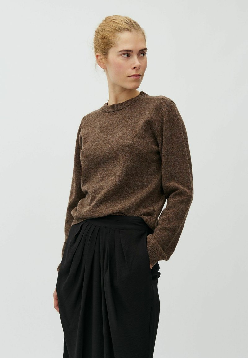 Femme aux cheveux blonds en chignon portant un pull marron à manches longues et une jupe plissée noire, regardant vers sa droite avec les mains dans les poches.