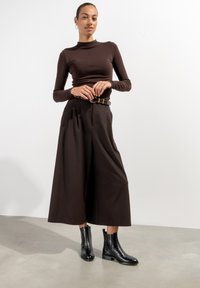 Brauner langärmeliger, taillierter Oberteil, kombiniert mit hochtaillierten, weit geschnittenen braunen Hosen und schwarzen Ankle Boots mit Krokodilstruktur und robustem Absatz.