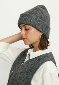 Woman wearing a gray knit beanie and matching buttoned vest over a white collared shirt, looking down with one arm raised behind her head.