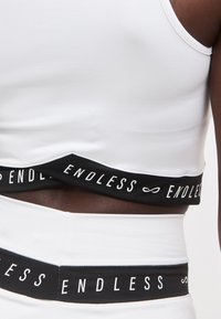 Εγγύς λήψη ατόμου που φοράει λευκό crop top και σορτς με μαύρες ελαστικές ταινίες που φέρουν την επιγραφή «ENDLESS» και σύμβολα απειροστικού.
