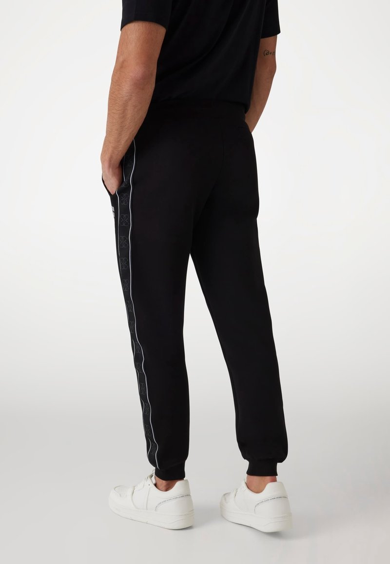 Schwarze Jogginghose mit schmal zulaufendem Schnitt, seitlichen Streifen mit Logo-Muster, elastischen Säumen und zwei Seitentaschen. Getragen mit weißen Sneakers.