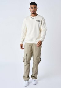 Sweatshirt blanc cassé avec le logo "PROJECT", associé à un pantalon cargo beige avec des poches latérales. Chaussures : baskets blanches.