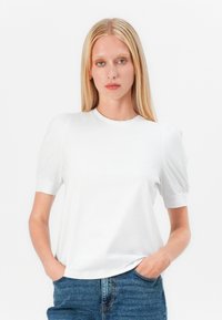 Blondínka v bielom tričku s balónovými rukávmi a modrých džínsach stojí s rukami v vreckách na bielom pozadí.