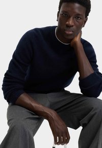 Joven sentado en un suéter azul marino y pantalones grises, descansando la cabeza sobre la mano, mirando directamente hacia adelante contra un fondo claro y sencillo.