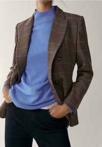 Blazer à carreaux marron avec des accents bleus sur un pull col roulé bleu. Le blazer présente des épaules structurées et des boutons double poitrine.