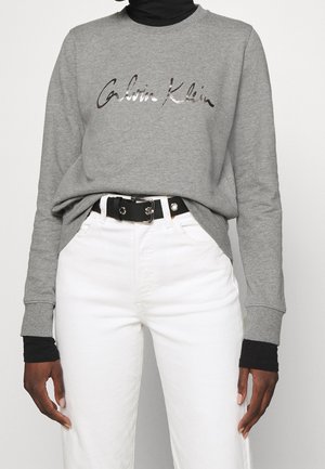 Person wearing grey Calvin Klein sweatshirt over black long sleeves, white high-waisted pants, and a black belt with silver buckle.