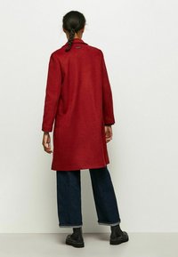 Czerwony długi płaszcz z kołnierzem, wykonany z teksturowanej tkaniny. Posiada długie rękawy, boczne kieszenie oraz prosty krój, zestawiony z ciemnymi spodniami dżinsowymi.