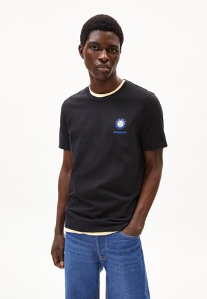 Maglietta nera in cotone con colletto girocollo a coste crema, decorata con una grafica di un sole blu e un logo sul petto. Indossata con jeans di denim blu.