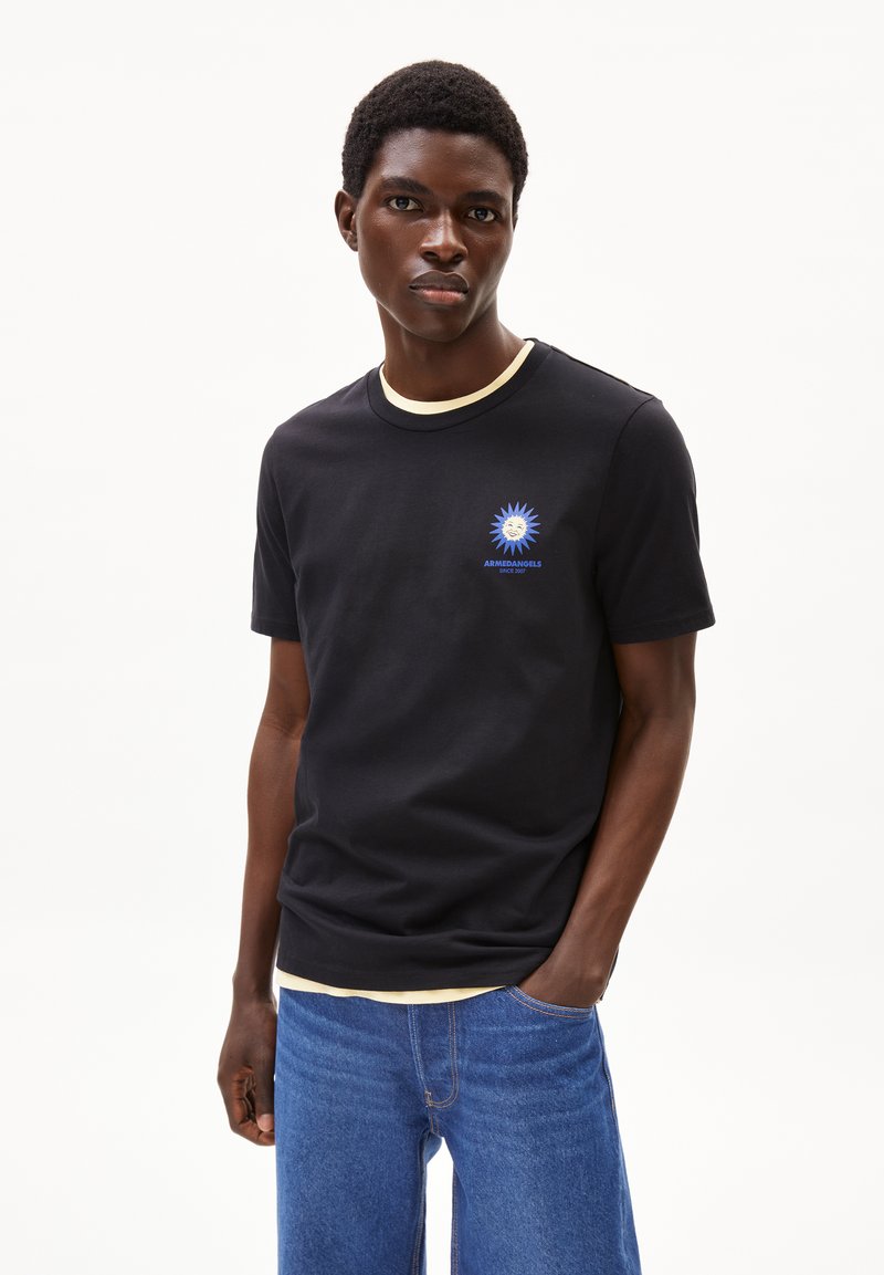 Maglietta nera in cotone con colletto girocollo a coste crema, decorata con una grafica di un sole blu e un logo sul petto. Indossata con jeans di denim blu.