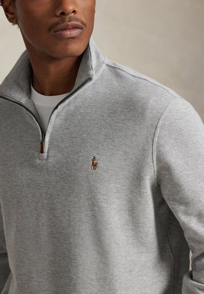 Homme portant un pull gris clair à zip quart avec petit logo brodé cheval et cavalier sur le côté gauche de la poitrine, zip ouvert sur une chemise blanche.