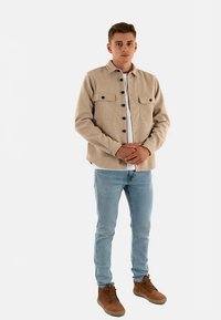 Beige jas met knopen en twee borstzakken, gedragen over een wit T-shirt, gecombineerd met lichtblauwe jeans en bruine veterlaarzen.