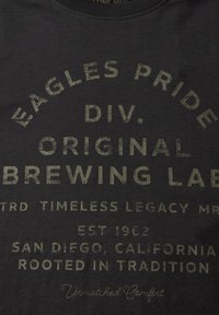 T-paita tummaa harmaata puuvillaa, jossa on suurta, kulutettua vihreää tekstiä. Teksti sisältää "EAGLES PRIDE", "ORIGINAL BREWING LAB" ja "SAN DIEGO, CALIFORNIA."