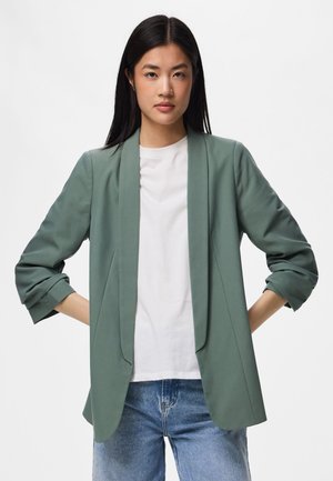 Femme debout face à l'objectif, les mains sur les hanches, portant un blazer vert, un t-shirt blanc et un jean bleu sur un fond blanc uni.