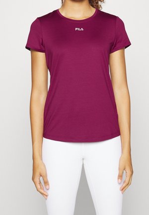 Kobieta w bordowej, krótkim rękawem koszulce Fila i białych spodniach, stojąca na tle jasnego, jednolitego tła.