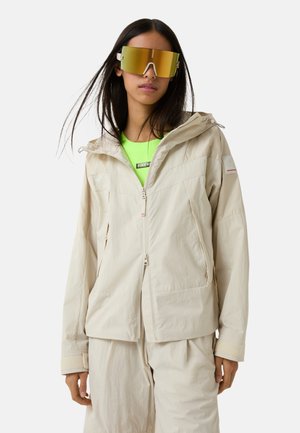 Beige lightweight jacket with a hood, featuring two front pockets, zipper closure, and a textured fabric. Worn over a bright green top.