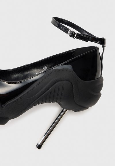 Pantofii negri din piele patentată cu toc înalt, având o curea la gleznă, și o talpă din cauciuc texturat negru, cu un toc stiletto metalic argintiu.