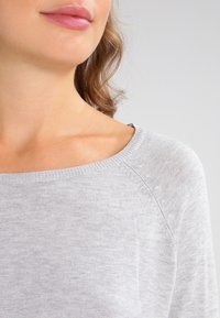 Jasnoszary sweter dzianinowy z szerokim dekoltem, prążkowanymi mankietami i miękką fakturą, w luźnym kroju z subtelnymi detalami szycia.