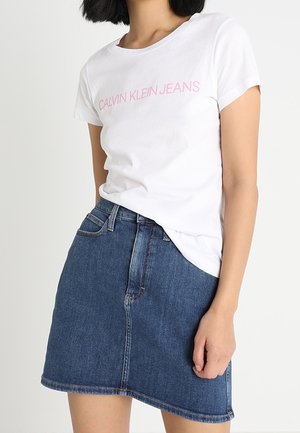 Kobieta w białej koszulce Calvin Klein Jeans i niebieskiej dżinsowej spódnicy do kolan, stojąca na jednolitym jasnym tle.