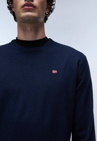 Marineblaue Strickjacke mit Rundhalsausschnitt, ausgestattet mit einer kleinen gestickten norwegischen Flagge auf der linken Brust. Glatte Textur.