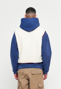 Fleece-lined hoodie with a soft white back, dark blue sleeves, and an adjustable hood. Features a ribbed hem and contrasting maroon accents.