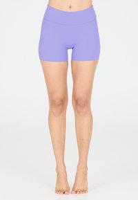 Nackte Beine vom Taille bis zu den Füßen, in engen, lila Sportshorts, stehend auf weißem Hintergrund.