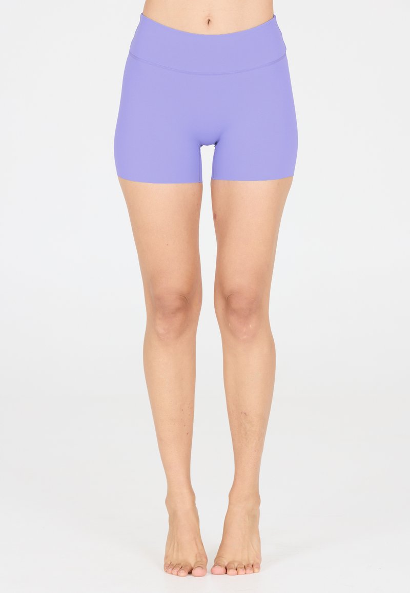 Nackte Beine vom Taille bis zu den Füßen, in engen, lila Sportshorts, stehend auf weißem Hintergrund.