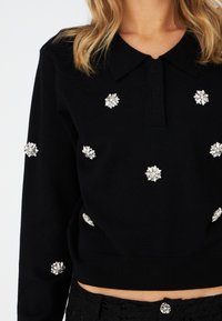 Maglione nero a maglia con colletto, caratterizzato da decorazioni floreali in argento distribuite uniformemente sul tessuto. Texture morbida, design aderente.