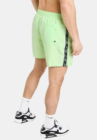 Pantaloni da nuoto verdi con tasca a toppa, vita elasticizzata e striscia laterale nera con branding bianco. Indossati con sneaker nere e bianche.